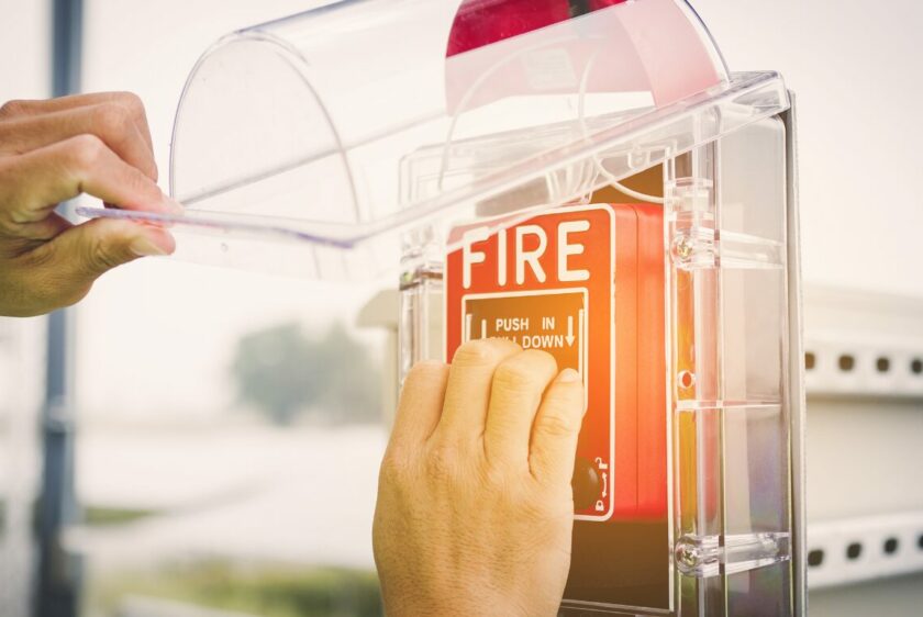 a man holding up the clear, plastic protection cover and activating the red fire alarm by pulling down the lever.