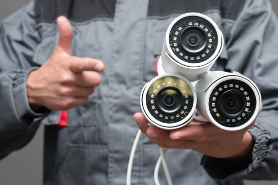 A RACAM security professional in a uniform holding three commercial CCTV security cameras while giving a thumbs-up.