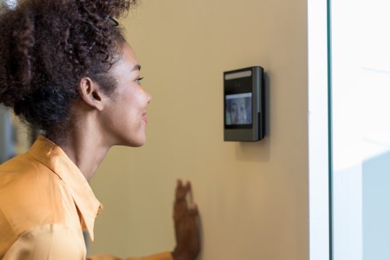 A women interacting with a facial recognition, video intercom system on a wall to gain access to the next room.